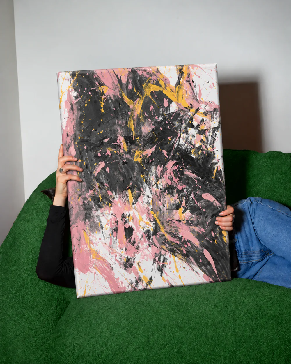 Osoba na zielonej kanapie trzyma przed sobą różowo-czarny obraz na ramie 120 × 90 cm